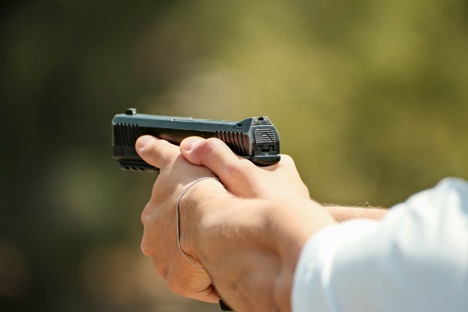 Close-up of a person's hands holding a black semi-automatic handgun, with a blurred outdoor background.